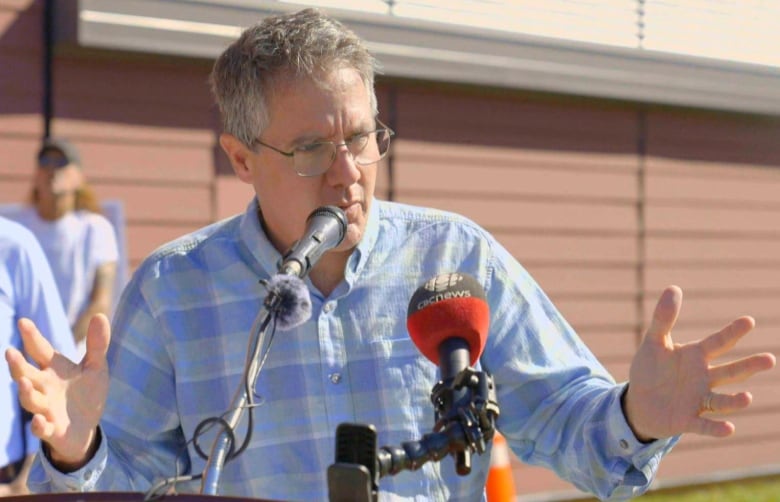 A grey-haired man in a blue-checked shirt stands behind a podium of microphones talking.