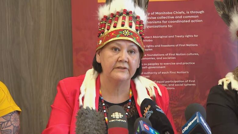 A woman wears a First Nations headdress and sits at a table with microphones.