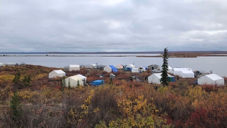 Wall tents and tarps sit amid autumn foliage on the shore of a lake.