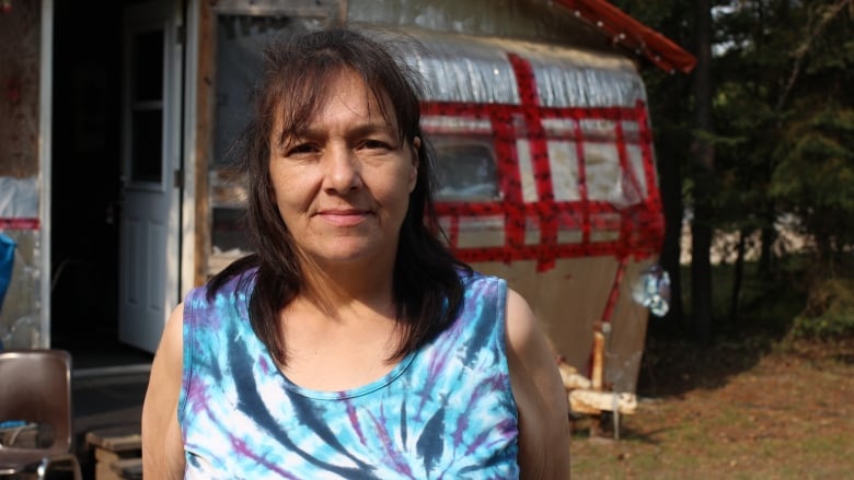 A woman in a tie-dyed shirt with brown hair stands in front of a trailer in Conklin, Alta.