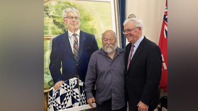 Two men smile as they stand next to a portrait showing one of the men.