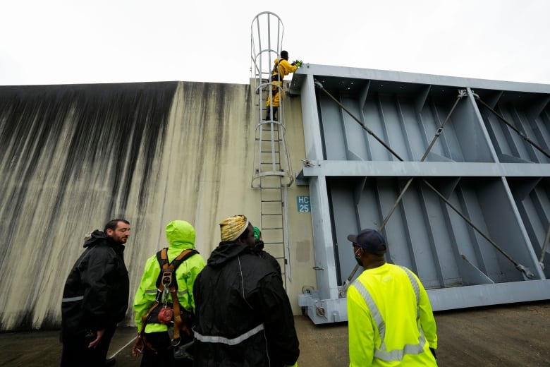 Four people stand in front of a tall concrete wall as another person stands atop a ladder inspecting a large steel floodgate. 