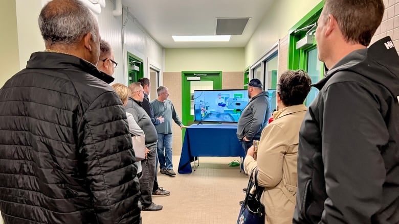 A small group of people stand in a hallway looking at a TV screen.