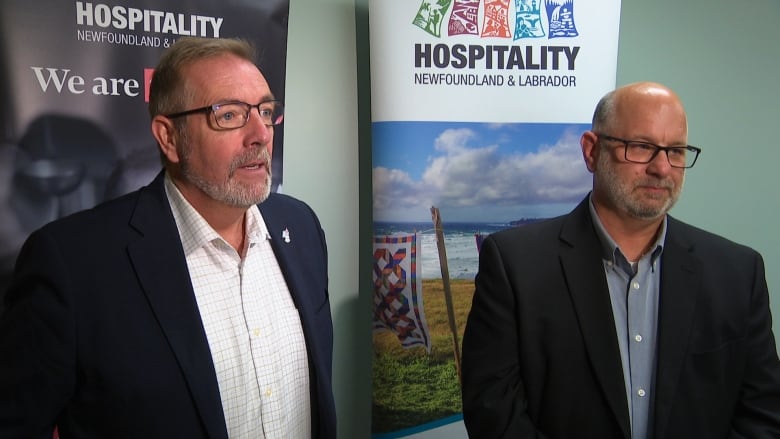 Two men wearing suits stand in front of banners for Hospitality Newfoundland and Labrador.