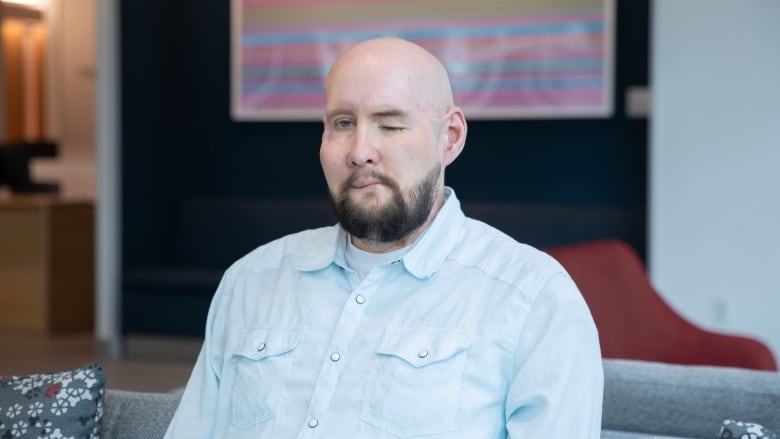 A bald man with a beard sits on a sofa and looks at the camera. The left side of his mouth curves upward, and his left eye is closed. 