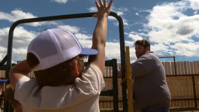 A man leans on a fence. In the foreground of the photo, a young child raises their hand.