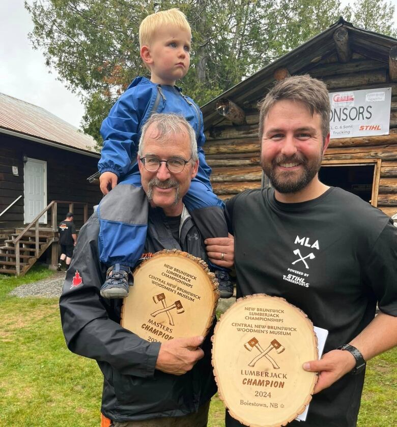 Two men standing next to each other and holding wooden plaques. A small child sits on the left man's shoulders.