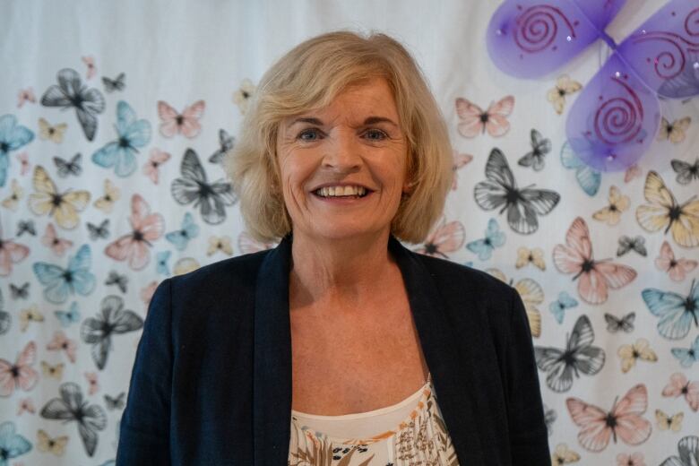 A person wearing a blazer is seen smiling in front of a butterfly-patterned wall.