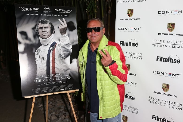 A man in sunglasses and a bright coloured jacket stands and makes a gesture nexts to a poster.