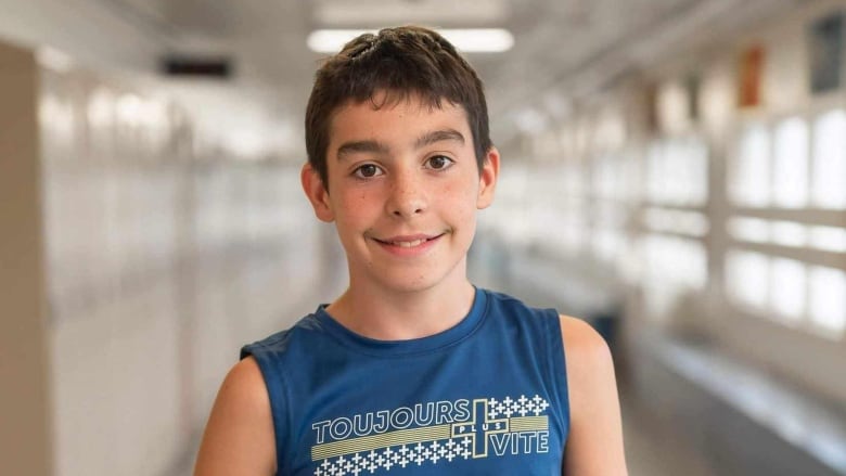 A picture of a boy smiling at the camera. Behind him is a school hallway.