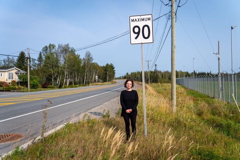 A woman stands next to a highway, in front of a speed limit sign 