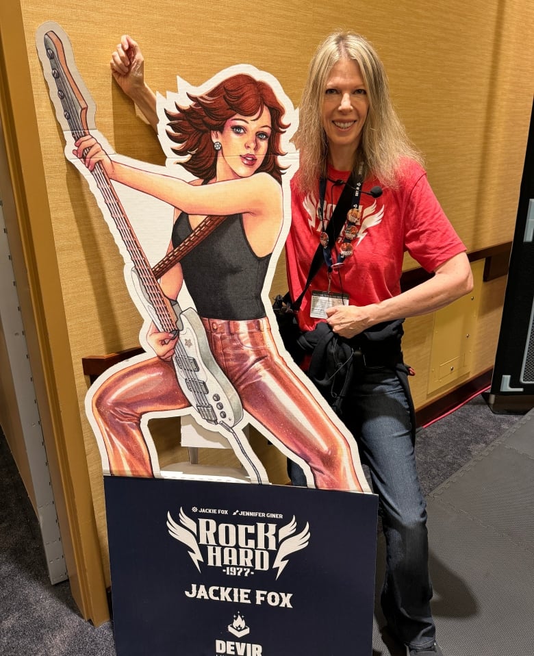 A smiling woman with shoulder-length blonde hair stands beside a life-sized cardboard cut-out that looks like a cartoon version of her younger self as the bassist for The Runaways. 