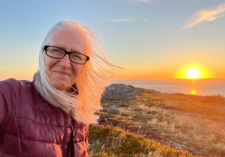 A woman takes a selfie on a path outside overlooking a body of water, with the sunset in the background.