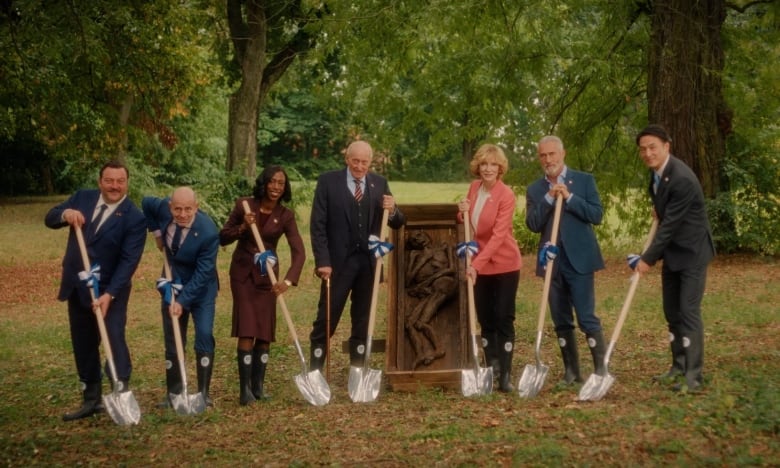 A publicity still shows seven people in suits holding shovels, with a coffin containing a skeletal body between them.