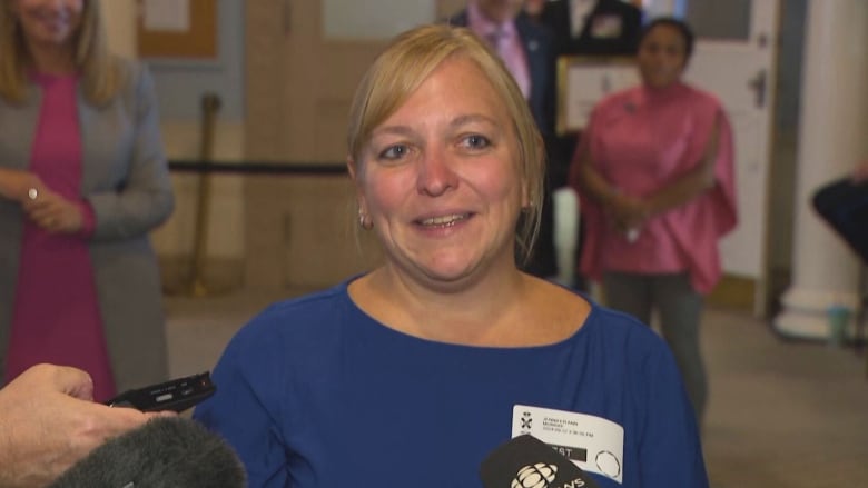 woman in blue shirt stands in front of microphones.