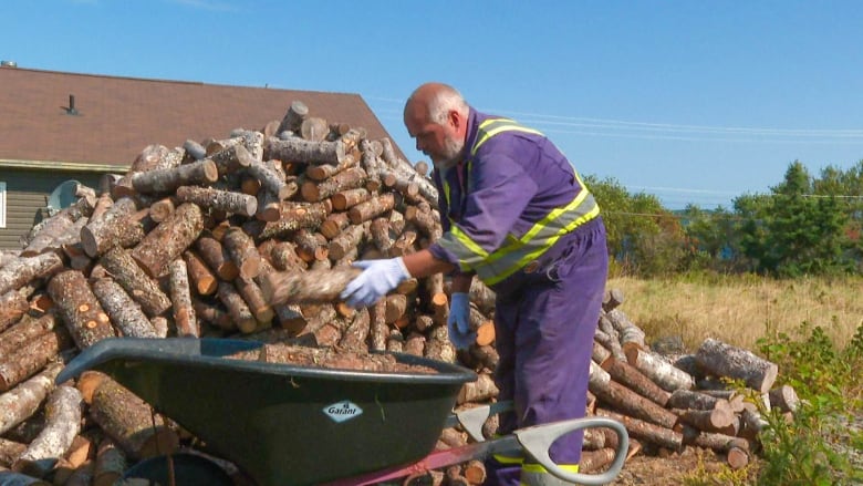 A bearded man with white gloves and coveralls, stacks wood into a green wheelbarrow