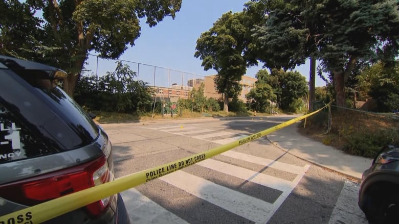 Yellow police tape blocks off a crime scene on a road with a school building in the background.