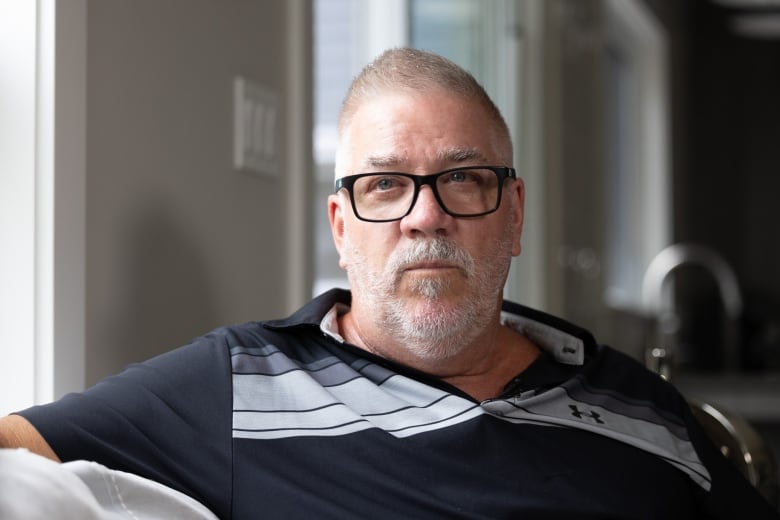 A Caucasian man with short hair and glasses sits on a couch, looking into the distance.