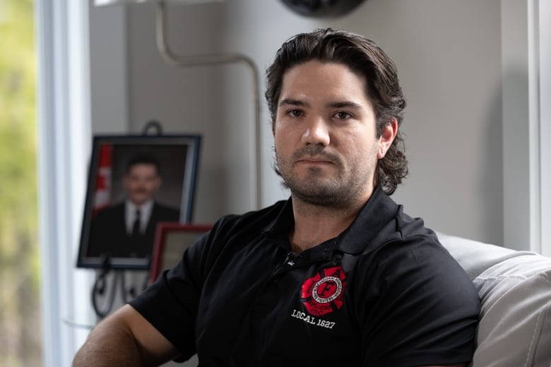 A firefighter with brown hair sits on a couch wearing a black shirt with a firefighters union logo on it. 