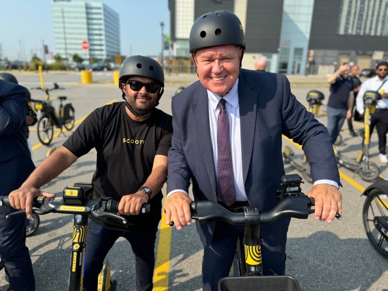 Two men wearing helmets pose with e-scooters.