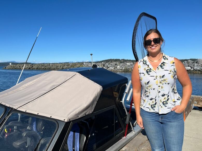 A woman in a sleveless flowered shirt stands on the dock beside her boat. The breakwater beneath the cloudless sky gives the impression of a nice day.