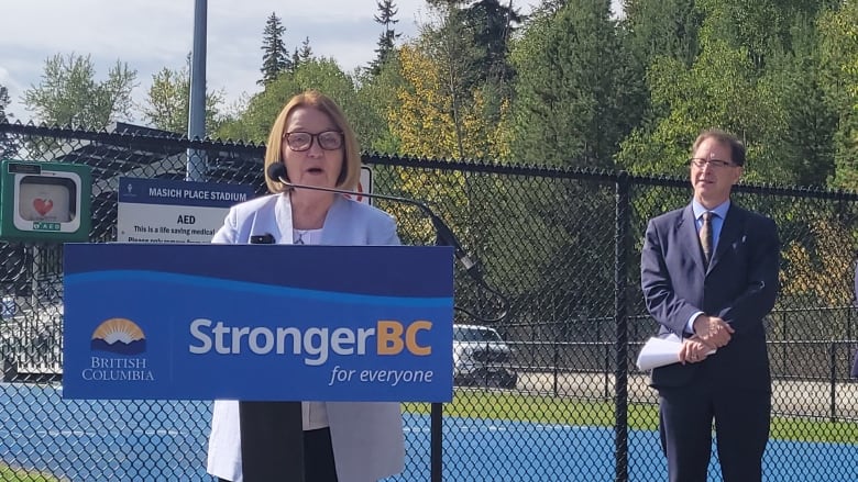 A woman at a podium as a man looks on.