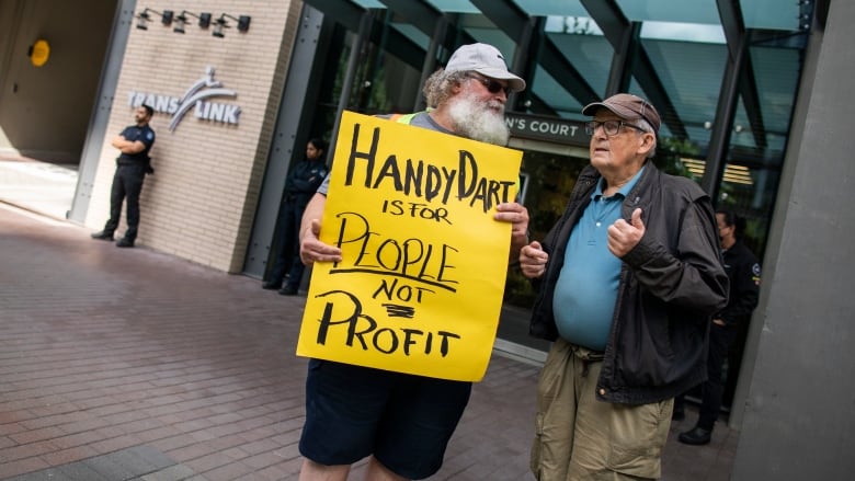 Two older men stand outside a corporate building, one of whom is holding a sign that reads 'HandyDART is for people, not profit'.