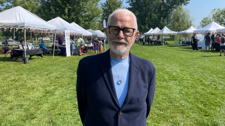 A man with white hair and white beard wearing black-framed glasses, navy blazer, sky-blue shirt and a ring hanging from a chain around his neck poses outside. Behind him are several white tents. 