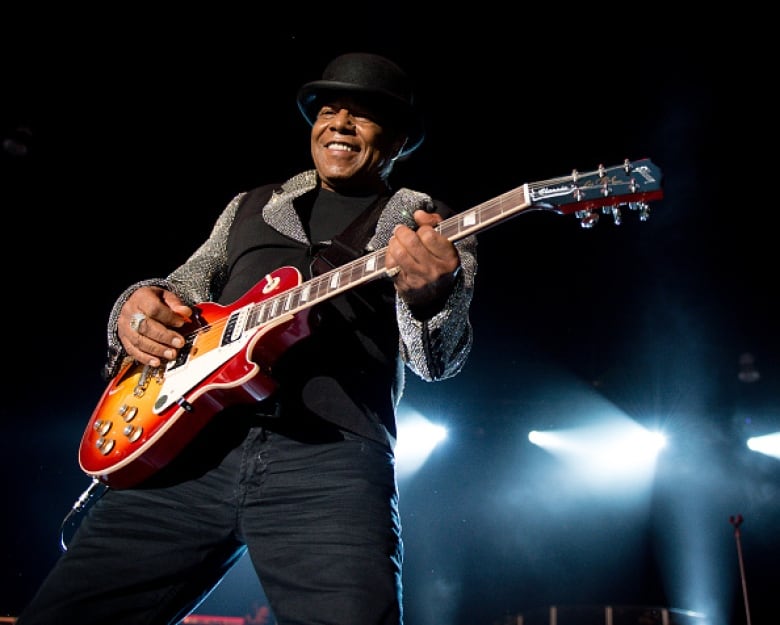 A man in a black vest and hat, smiles as he plays an electric guitar on stage.