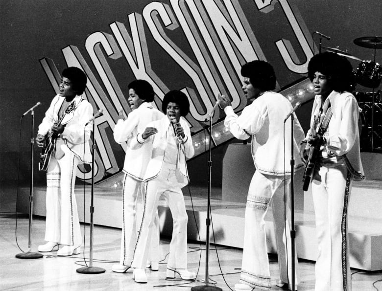 Five young men in white suits sing on a stage.