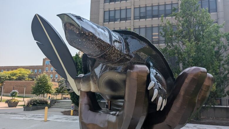 A metal sculpture of a hand emerging from the earth at the wrist,  The hand is tilted backwards with a turtle -- who is about the same size as the palm and fingers -- sitting inside it holding a feather.