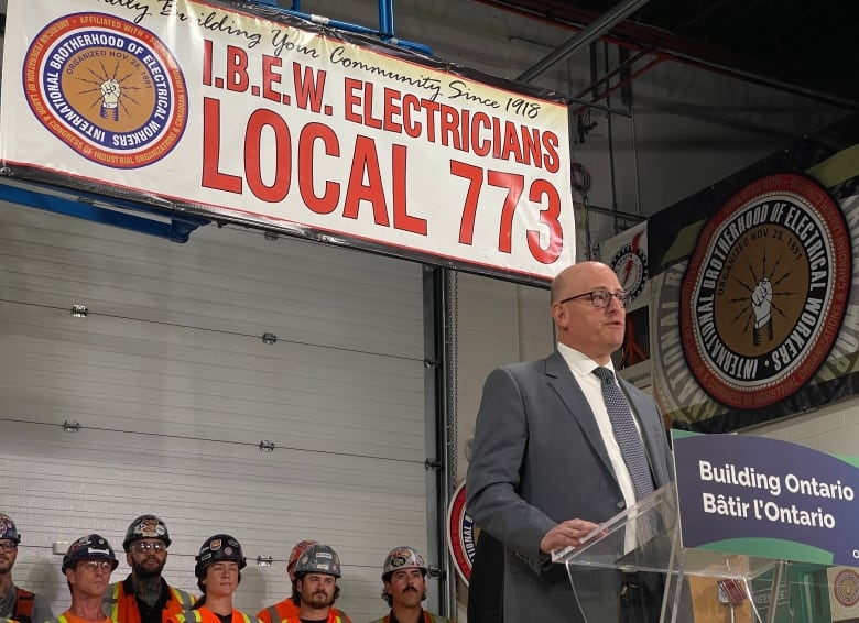 Drew Dilkens at a podium with a row of workers in hard hats and hi-viz vests standing behind him.