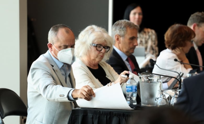 A man in a medical face mask picking up papers seated alongside others behind a desk with microphones and a pitcher of water.