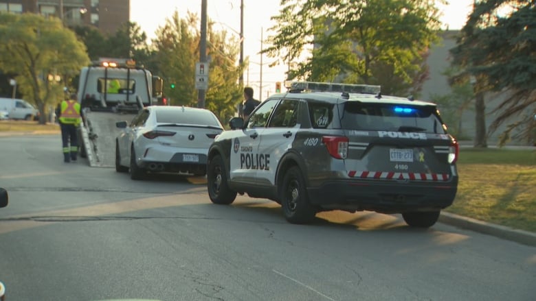 A police SUV with lights on is parked on a curb behind a white sedan that is about to be rolled up the ramp of a tow truck. It's late evening. Long shadows and sunlight break through trees in a residential area