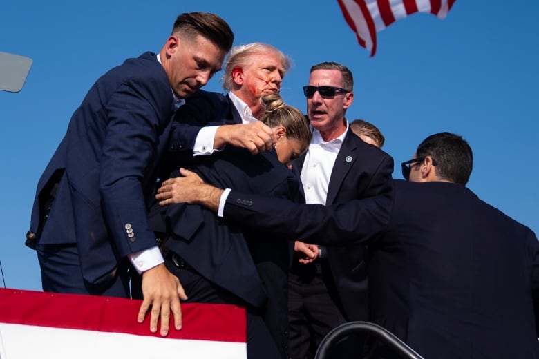 A group of secret service agents in suits and sunglasses surround a man who looks dazed, with a small trickle of blood down the side of his face.