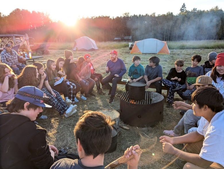 A big group of students sit around a fire outside during the day. 