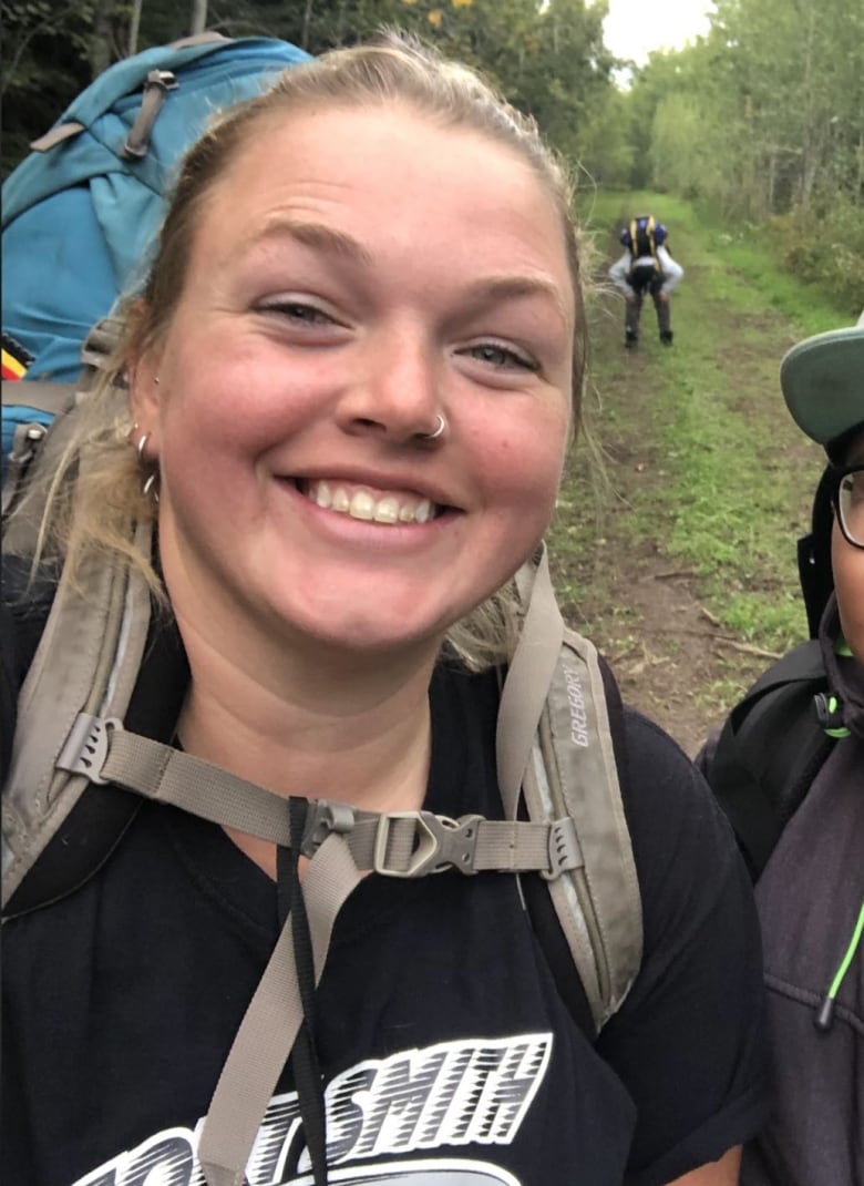 A blonde woman outside with a backpack on smiles at the camera 