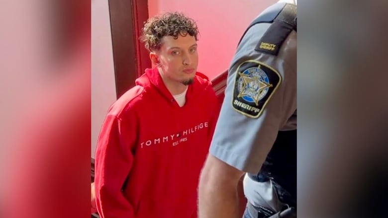 A man in a red hoodie looks toward the camera. The arm of a court sheriff is visible in the right side of the frame.