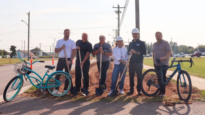 Six people holding shovels standing beside two bicycles.