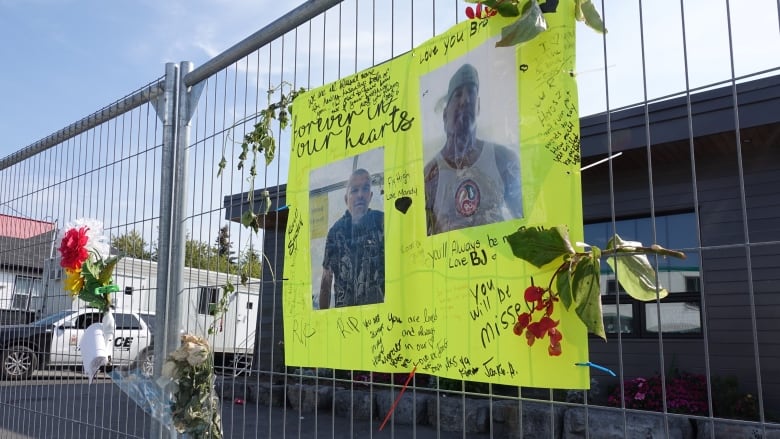 A lime green poster board with photos of two men is surrounded by messages of love and condolences. Bouquets of flowers are tied to the fence and a police cruiser is parked in the background.