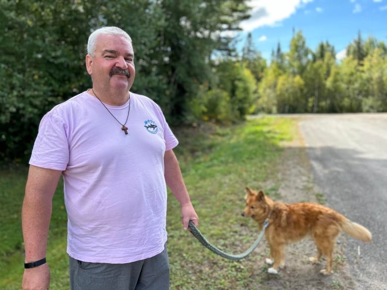 A man with grey hair smiles at the camera while holding a leash to his brown, mid-sized dog.