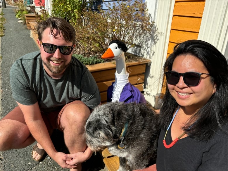 A man and a woman wearing sunglasses sit next to a goose statue that's dressed in a purple jacket and a wig that impersonates Prince.