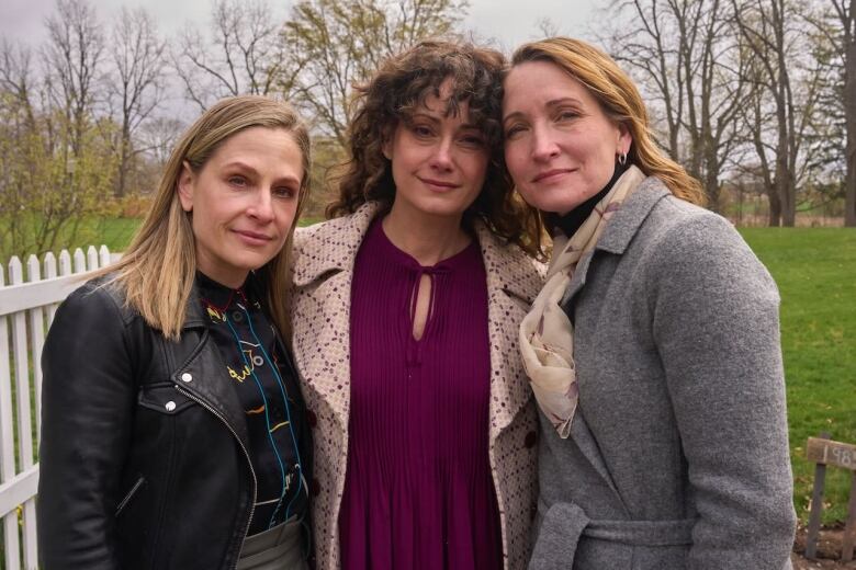 a blonde woman in a black leather jacket stands beside a dark-haired woman in the centre in a burgundy top and a white checkered coat, and at far right, is a blonde woman in a grey coat and a white scarf.