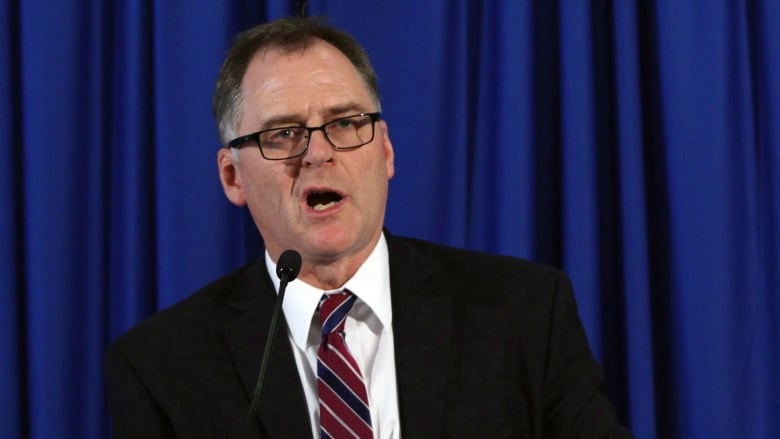 A man wearing a suit talks in front of a blue curtain.