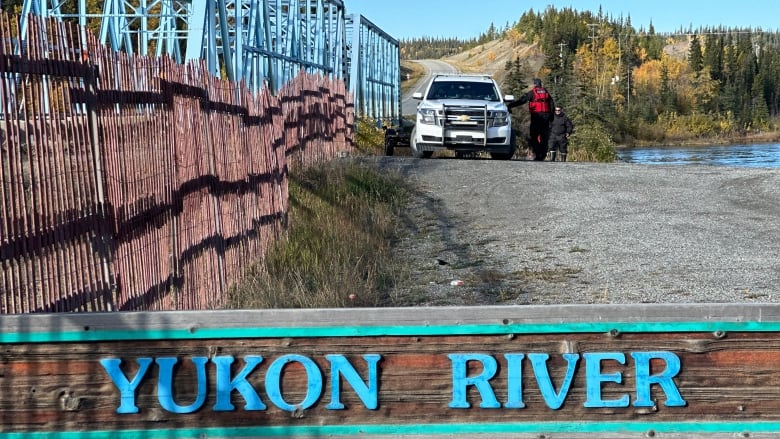 Two men standing next to a police vehicle on the banks of river next to a bridge. 