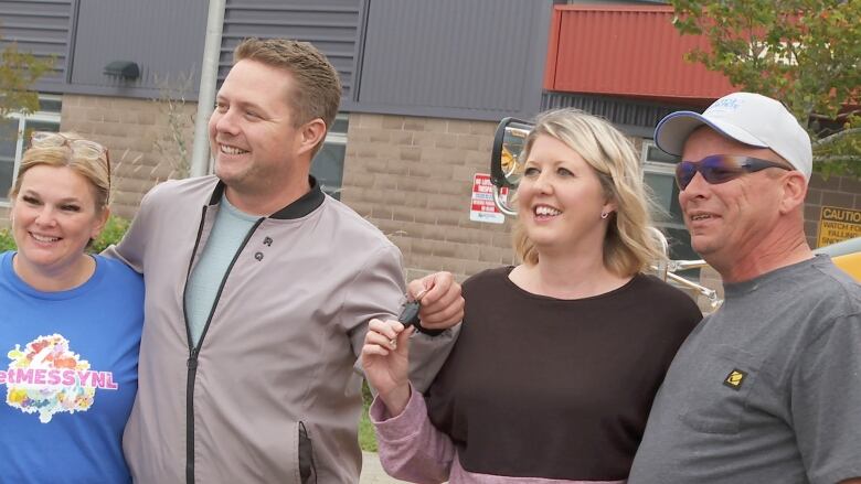 Four adults stand in a line smiling. A man and woman in the centre hold the key to a car.