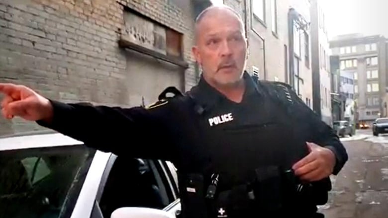 A police officer in uniform stands next to a car in an alley, with one arm pointed to the left.