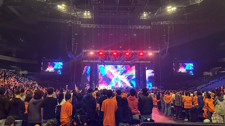 Students wearing orange shirts cheer in their seats for a group performing on stage. 