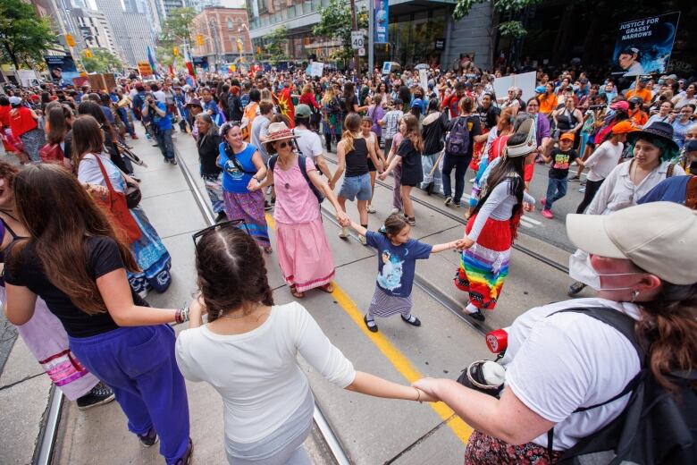 A large group of people gathers on a street. In the centre, a number of people are seen holding hands.