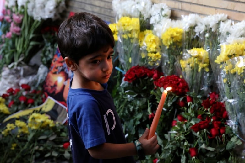 A young boy holds a lit candle in front of flowers on the ground.